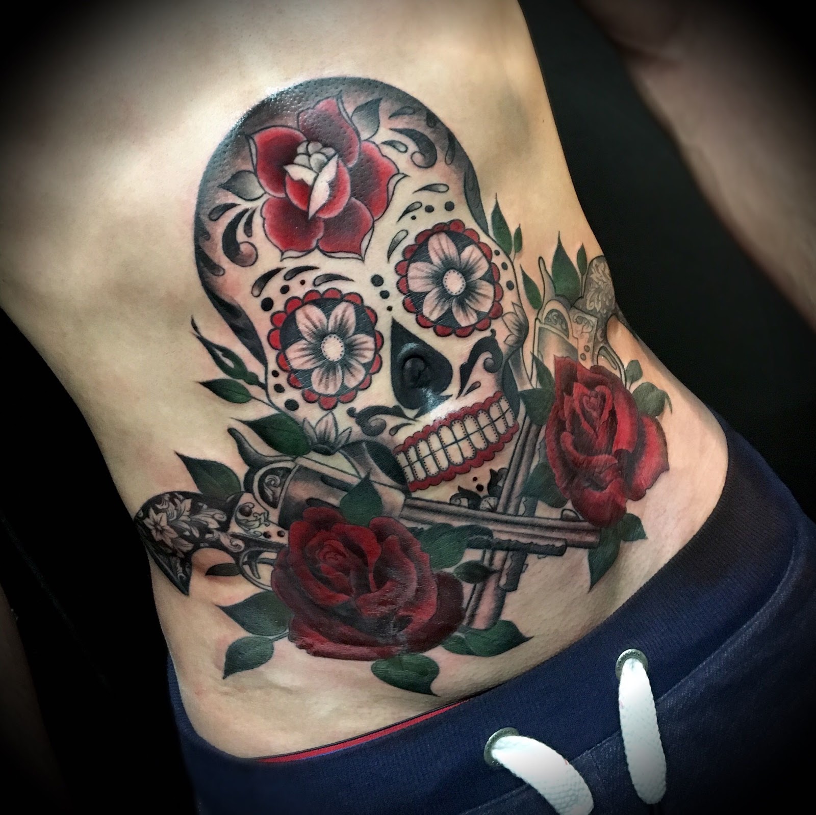 Sugar skull Día de los Muertos avec roses rouges et revolvers, tatouage couleur sur le flanc